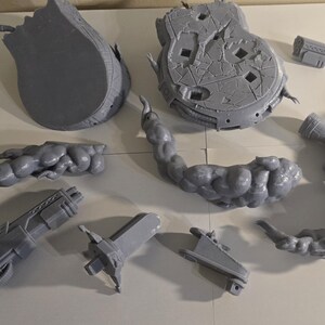 May include: A collection of gray, 3D-printed model parts, including a large, broken circular base, a curved, cloud-like structure, and various mechanical components. The pieces are arranged on a white surface.