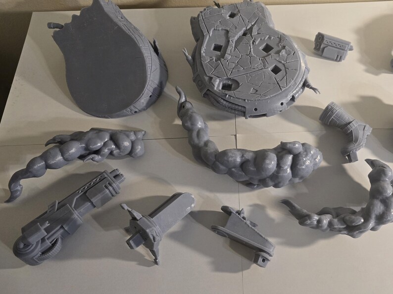 May include: A collection of gray, 3D-printed model parts, including a large, broken circular base, a curved, cloud-like structure, and various mechanical components. The pieces are arranged on a white surface.
