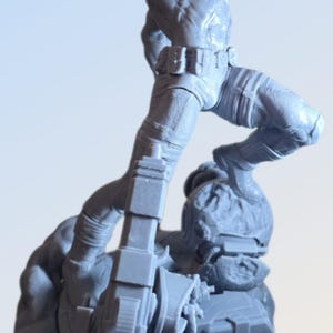 May include: A gray action figure of a muscular character in a dynamic pose, with one leg raised and arm overhead. The figure is atop a detailed base with mechanical elements. The figure has a red element on its face.