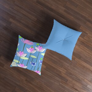 May include: Two square throw pillows. One pillow features a light blue background with a pattern of pink and purple lotus flowers. The other pillow is solid light blue with a button in the center.