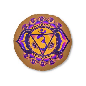 May include: Round brown cushion featuring a vibrant chakra design. The design includes a central yellow triangle with a purple Om symbol, surrounded by intricate purple and yellow floral patterns. The cushion is a decorative item.
