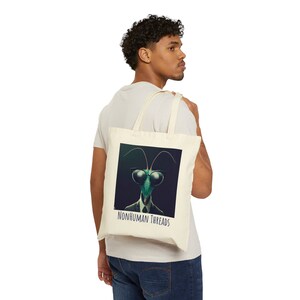 May include: Cream-colored tote bag featuring a graphic of a praying mantis wearing sunglasses and a suit. The text "NONHUMAN THREADS" is printed below the image. The bag is carried over a person's shoulder.
