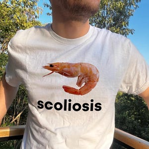 May include: White t-shirt with a cooked prawn graphic in orange and red. The word "scoliosis" is printed in black beneath the prawn. The shirt is being worn. The background is blurred.
