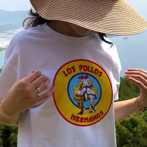 Peut inclure: Un t-shirt blanc avec un graphique circulaire jaune représentant deux poulets de dessin animé et le texte "Los Pollos Hermanos".