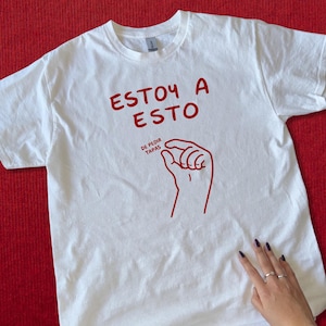 Könnte beinhalten: Weißes T-Shirt mit rotem Text und einer Handgrafik. Der Text lautet "ESTOY A ESTO" über der Hand und "DE PEDIR TAPAS" darunter. Das Shirt liegt auf rotem Untergrund.