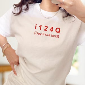 Pode incluir: T-shirt branca de manga curta com o texto vermelho "i 124 Q (Say it out loud)". A t-shirt é feita de um material macio.