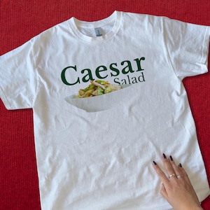Puede incluir: Camiseta blanca con las palabras "Caesar Salad" en verde y una imagen de una ensalada César. La camiseta está sobre una superficie roja. Una mano con anillos toca la camiseta.