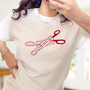 Op de afbeelding: Wit T-shirt met een rode schaar afbeelding. Het schaarontwerp is een gestileerde illustratie, gecentreerd op het T-shirt. Het T-shirt heeft een klassieke ronde hals.