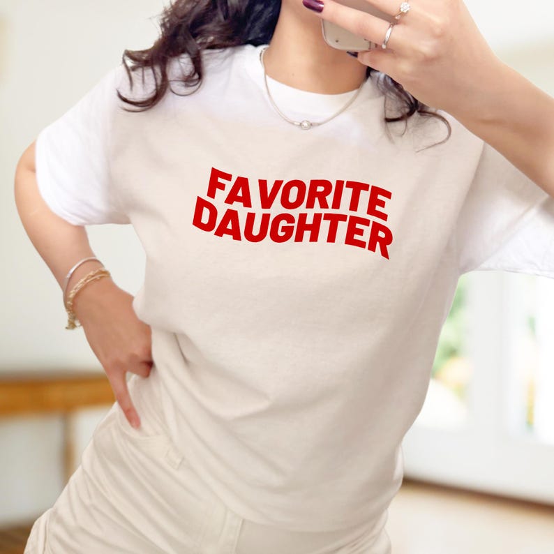 Puede incluir: Camiseta blanca con el texto rojo "FAVORITE DAUGHTER". La camiseta tiene mangas cortas y cuello redondo. La imagen muestra a una persona usando la camiseta, con joyas y un tel&eacute;fono.