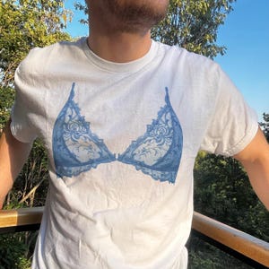 May include: White t-shirt featuring a blue lace bra design. The bra graphic has intricate floral patterns and lace detailing. The shirt is a crew neck style and appears to be made of a soft, comfortable material.