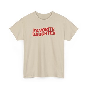 Puede incluir: Una camiseta beige con la frase "FAVORITE DAUGHTER" en letras rojas may&uacute;sculas. La camiseta tiene cuello redondo y est&aacute; hecha de una tela suave. Una prenda sencilla e informal para el uso diario.
