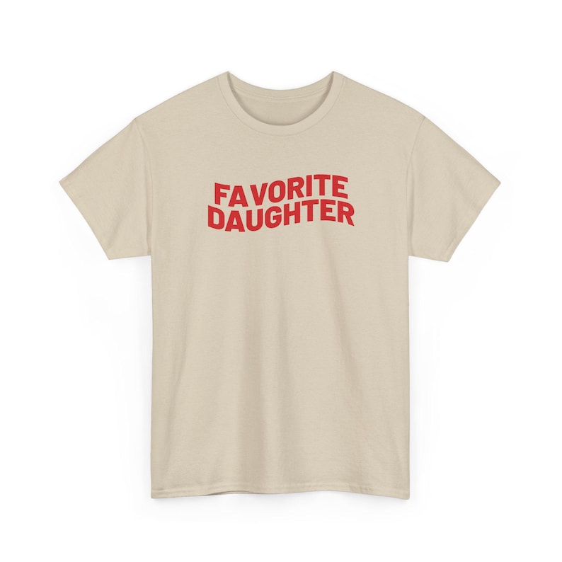 Puede incluir: Una camiseta beige con la frase "FAVORITE DAUGHTER" en letras rojas may&uacute;sculas. La camiseta tiene cuello redondo y est&aacute; hecha de una tela suave. Una prenda sencilla e informal para el uso diario.
