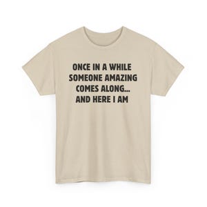 Op de afbeelding: Lichtbeige t-shirt met zwarte tekst: "ONCE IN A WHILE SOMEONE AMAZING COMES ALONG... AND HERE I AM". Het t-shirt heeft korte mouwen en een ronde hals.
