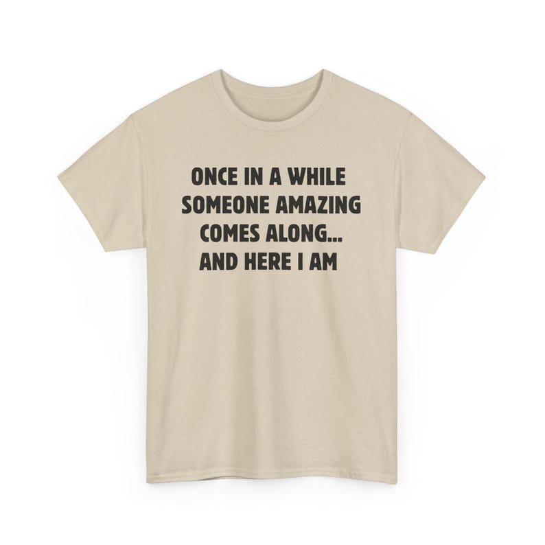 Op de afbeelding: Lichtbeige t-shirt met zwarte tekst: "ONCE IN A WHILE SOMEONE AMAZING COMES ALONG... AND HERE I AM". Het t-shirt heeft korte mouwen en een ronde hals.