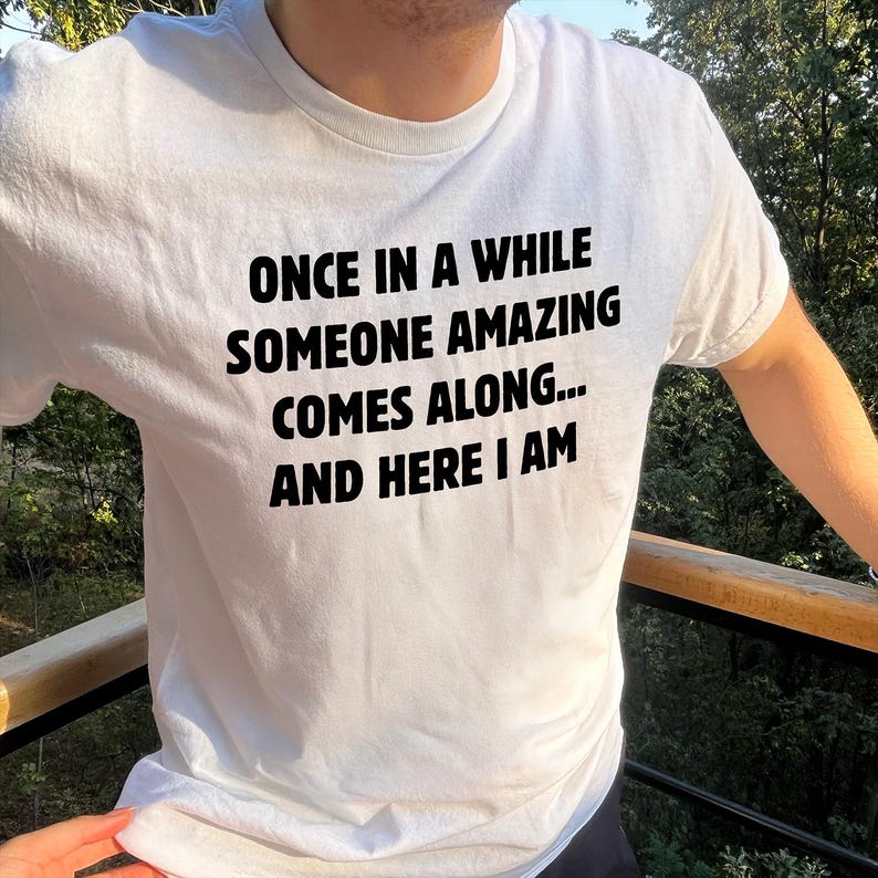 Op de afbeelding: Wit T-shirt met zwarte tekst: "ONCE IN A WHILE SOMEONE AMAZING COMES ALONG... AND HERE I AM". Het T-shirt heeft een ronde hals. De foto is buiten genomen, met groen op de achtergrond.