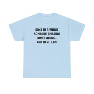 Op de afbeelding: Lichtblauw T-shirt met de zwarte tekst: "ONCE IN A WHILE SOMEONE AMAZING COMES ALONG... AND HERE I AM". Het shirt heeft een ronde hals en korte mouwen. De tekst staat in het midden op de voorkant.