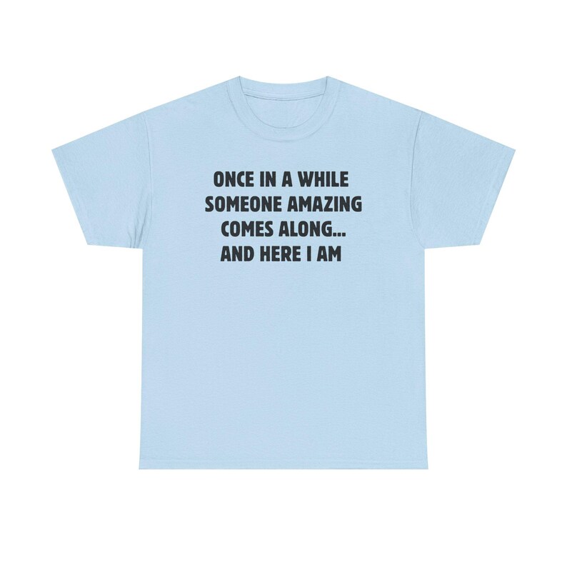 Op de afbeelding: Lichtblauw T-shirt met de zwarte tekst: "ONCE IN A WHILE SOMEONE AMAZING COMES ALONG... AND HERE I AM". Het shirt heeft een ronde hals en korte mouwen. De tekst staat in het midden op de voorkant.