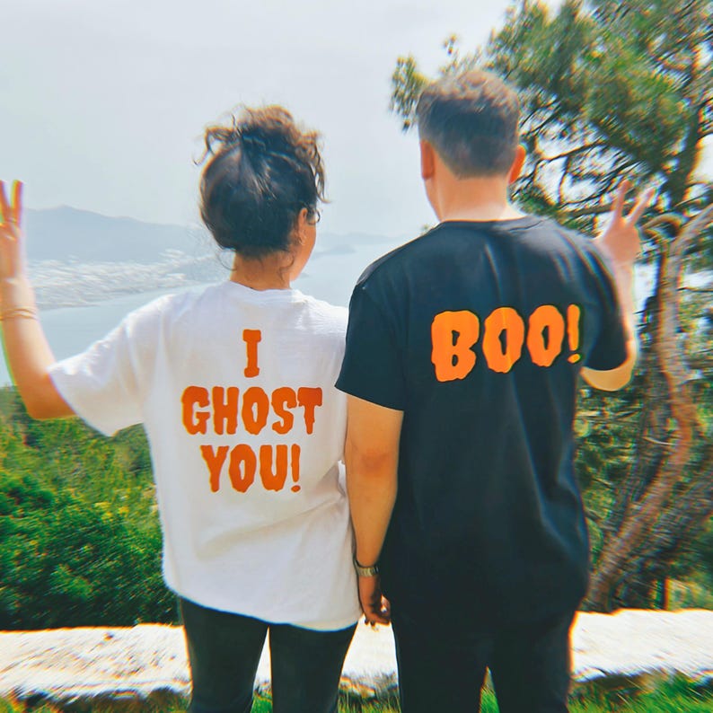 May include: Two people wearing matching Halloween themed shirts. The person on the left is wearing a white shirt with the text "I Ghost You!" in orange. The person on the right is wearing a black shirt with the text "BOO!" in orange.