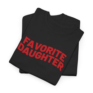 Puede incluir: Dos camisetas negras apiladas, con la camiseta superior que muestra el texto rojo "FAVORITE DAUGHTER". Las camisetas est&aacute;n dobladas cuidadosamente, con el texto en un lugar destacado. El fondo es blanco liso.