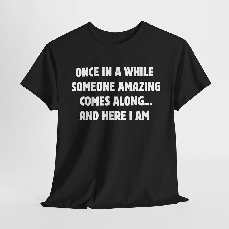 Op de afbeelding: Zwart T-shirt met witte tekst: "ONCE IN A WHILE SOMEONE AMAZING COMES ALONG... AND HERE I AM". De tekst staat in witte letters op de voorkant. Het shirt heeft korte mouwen en een ronde hals.