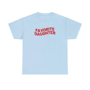 Puede incluir: Camiseta azul claro con la frase "FAVORITE DAUGHTER" en rojo en el pecho. El texto est&aacute; en una fuente audaz y juguetona. Es una prenda casual para el d&iacute;a a d&iacute;a.
