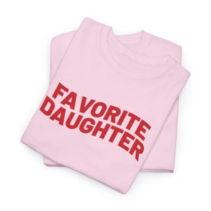 Puede incluir: Dos camisetas de color rosa claro apiladas una encima de la otra. La camiseta superior tiene las palabras "FAVORITE DAUGHTER" impresas en rojo, arqueadas en la parte delantera. Las camisetas est&aacute;n dobladas cuidadosamente.