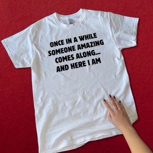 Op de afbeelding: Wit T-shirt met de tekst "ONCE IN A WHILE SOMEONE AMAZING COMES ALONG... AND HERE I AM" in het zwart. Het shirt wordt gepresenteerd op een rode ondergrond. Het shirt heeft een ronde hals.