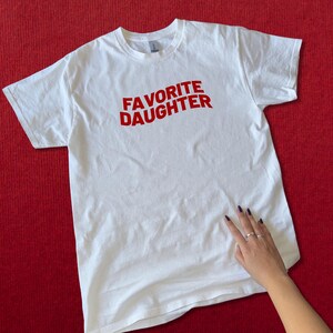 Puede incluir: Camiseta blanca con la frase "FAVORITE DAUGHTER" en rojo arqueada en la parte delantera. La camiseta est&aacute; sobre una superficie roja. Una mano con u&ntilde;as oscuras y anillos toca la camiseta.
