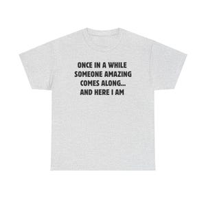 Op de afbeelding: Lichtgrijs t-shirt met de zwarte tekst: "ONCE IN A WHILE SOMEONE AMAZING COMES ALONG... AND HERE I AM". Het shirt heeft een ronde hals en korte mouwen. De stof lijkt een zachte katoenmix te zijn.