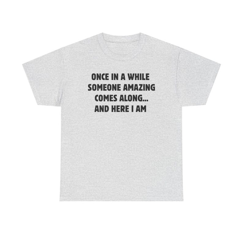 Op de afbeelding: Lichtgrijs t-shirt met de zwarte tekst: "ONCE IN A WHILE SOMEONE AMAZING COMES ALONG... AND HERE I AM". Het shirt heeft een ronde hals en korte mouwen. De stof lijkt een zachte katoenmix te zijn.