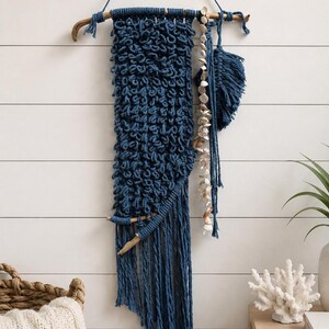 May include: A dark blue macrame wall hanging suspended from a horizontal piece of driftwood. The piece features a dense, knotted texture, a long fringe, a vertical strand of small white seashells, and a separate blue fiber leaf-shaped element.
