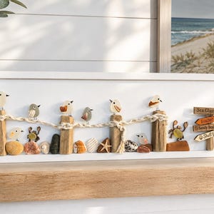 May include: A rectangular white wall art piece featuring pebble art birds perched on driftwood posts connected by rope. The scene includes small shells, stones, a starfish, and a crab. A wooden signpost displays the text Sea Glass Pier, Feather Cove, Sandy Beaks Club, and Shell Yeah!
