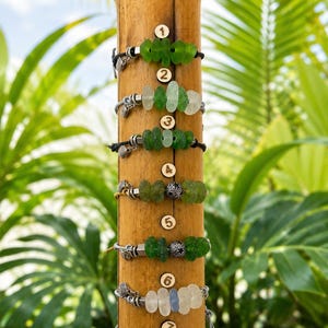May include: Seven bracelets displayed on a vertical bamboo stand, each numbered one through seven. The bracelets feature various sea glass pieces in shades of green, white, and blue, accented with small silver-toned metal charms and beads.