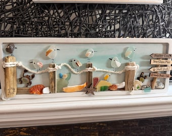 May include: A rectangular wall art piece featuring sea glass birds perched on driftwood posts connected by rope. The scene includes shells, a starfish, and a signpost with the text Feather Cove, Sea Glass Pier, Shell Yeah!, and Sandy Beaks Club.