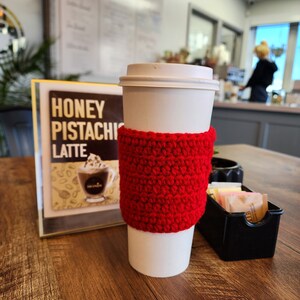 Könnte beinhalten: Ein weißer Einweg-Kaffeebecher mit einer leuchtend roten, strukturierten Häkelmanschette. Der Becher steht neben einem Schild mit der Aufschrift Honey Pistachio Latte und einem kleinen schwarzen Behälter mit verschiedenen Zucker- und Süßstoffpäckchen.