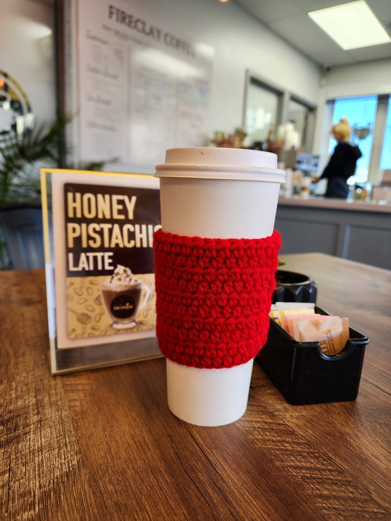 May include: A white disposable coffee cup featuring a bright red, textured crochet sleeve. The cup is positioned next to a sign that reads Honey Pistachio Latte and a small black container filled with various packets of sugar and sweeteners.