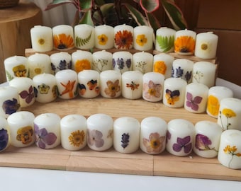 May include: A collection of white pillar candles, each adorned with a pressed flower design. The flowers include sunflowers, orange blooms, and purple pansies. The candles are arranged on a wooden surface.