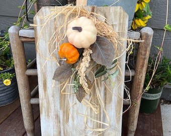 May include: A vertical wooden board shaped like a pumpkin with a light brown stem, decorated with a cluster of raffia, a small cream-colored pumpkin, a small orange pumpkin, and several brown and green artificial leaves and dried floral accents.