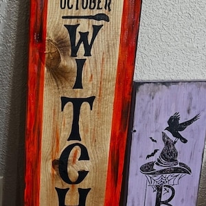May include: Two vertical wooden signs. The taller sign is natural wood with red painted edges, featuring a black witch hat, the text IT'S OCTOBER WITCHES, and witch boots. The shorter sign is light purple with black graphics of a hat, crow, and BOO.