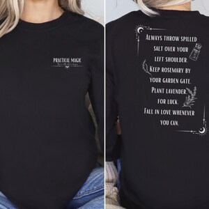 Könnte beinhalten: Schwarzes Langarm-Sweatshirt mit weißem Text. Auf der Vorderseite steht "PRACTICAL MAGIC". Auf der Rückseite befindet sich eine Liste mit Anweisungen und Illustrationen. Der Text enthält Sätze wie "verschüttetes Salz werfen" und "Lavendel pflanzen".