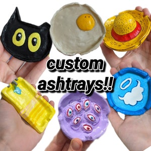 Puede incluir: Seis ceniceros personalizados hechos de arcilla con varios diseños. Los diseños incluyen un gato negro, un huevo frito, un sombrero de paja, una piscina, un cenicero morado con múltiples ojos y un cenicero azul con una nube y un halo.