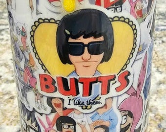 May include: A white tumbler covered in cartoon images and text from the animated series Bob's Burgers. The tumbler features the character Tina Belcher with the word "BUTTS" and the phrase "I like them."