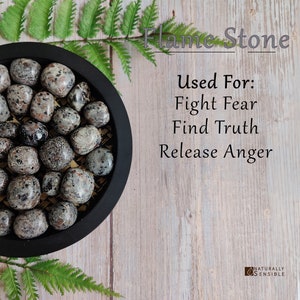 May include: A collection of black and white speckled tumbled stones, known as Flame Stone, in a black bowl. The stones are used for fighting fear, finding truth, and releasing anger. The text "Flame Stone" and "Used For: Fight Fear Find Truth Release Anger" is visible in the image.