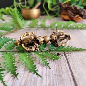 Puede incluir: Un objeto ritual vajra de color oro metálico con intrincadas tallas simétricas en ambos extremos, un agarre esférico central y puntas ornamentadas que se curvan hacia adentro para encontrarse en un extremo puntiagudo.