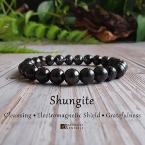 May include: A black Shungite bracelet with round beads. The bracelet is on a wooden surface. The text "Shungite" is written in white letters below the bracelet. The text "Cleansing • Electromagnetic Shield •  Gratefulness" is written below the word "Shungite". The text "Naturally Sensible" is written in white letters above the bracelet.