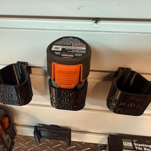 May include: Black 3D-printed battery holders and a power tool battery. The holders are labeled "for RIDGID HYPER". The power tool battery is black and orange. The items are mounted on a white slatted wall.