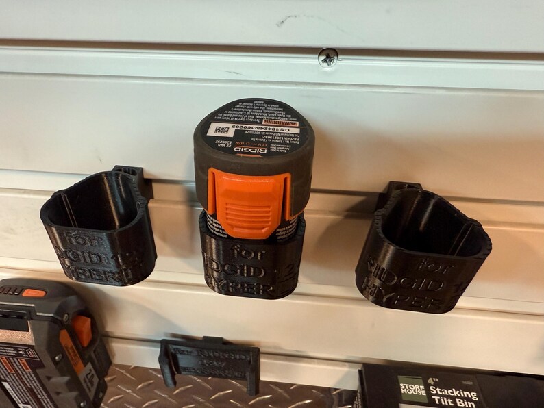 May include: Black 3D-printed battery holders and a power tool battery. The holders are labeled "for RIDGID HYPER". The power tool battery is black and orange. The items are mounted on a white slatted wall.
