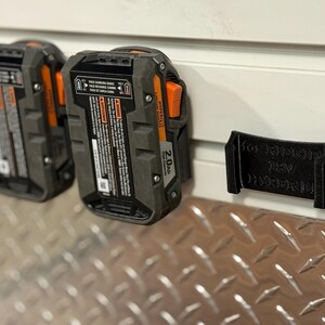 Puede incluir: Dos bater&iacute;as de herramientas el&eacute;ctricas de 18 V negras y grises con detalles en naranja est&aacute;n montadas en una pared blanca. Un soporte negro para bater&iacute;as tambi&eacute;n est&aacute; montado en la pared. Las bater&iacute;as son de 2,0 Ah.