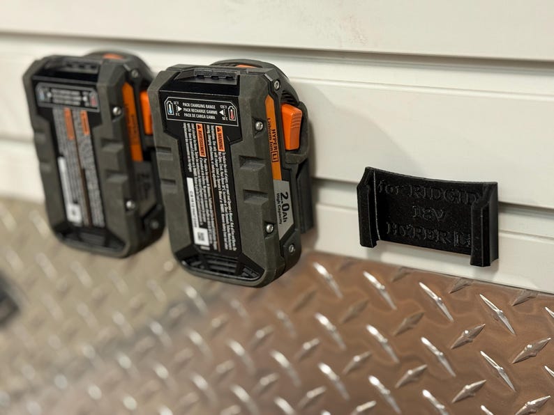 Puede incluir: Dos bater&iacute;as de herramientas el&eacute;ctricas de 18 V negras y grises con detalles en naranja est&aacute;n montadas en una pared blanca. Un soporte negro para bater&iacute;as tambi&eacute;n est&aacute; montado en la pared. Las bater&iacute;as son de 2,0 Ah.