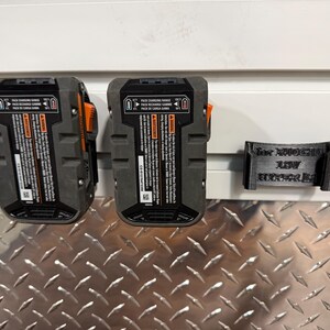 Puede incluir: Dos bater&iacute;as de herramientas el&eacute;ctricas negras y grises con detalles en naranja est&aacute;n montadas en una pared, junto a un soporte de bater&iacute;a negro impreso en 3D. Las bater&iacute;as tienen etiquetas de advertencia e informaci&oacute;n del producto. El soporte tiene texto.